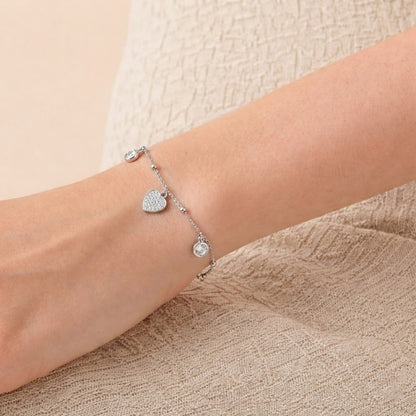 Silver bracelet with heart and round charm on a wrist against a beige background