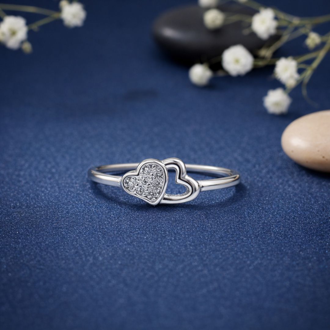 Silver heart-shaped ring with embedded stones on a blue textured surface with white flowers.